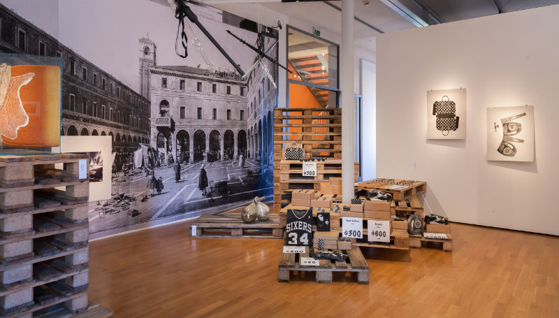 Blick in die Ausstellung mit Kunst auf Europaletten, Fototapete und an der Wand hängenden Drucken