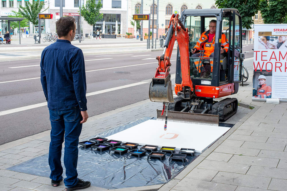 ein Mann in blauer Kleidung steht vor einem Minibgger, an den ein Pinsel montiert ist, mit dem er auf eine große weiße Fläche am Boden ein Bild malt