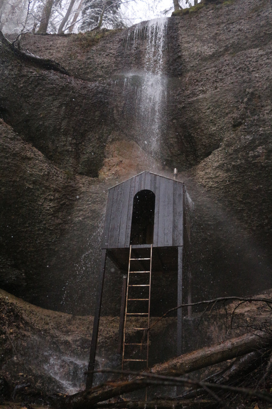 eine Holzhütte steht auf Stelzen unter einem Wasserfall am Felsen