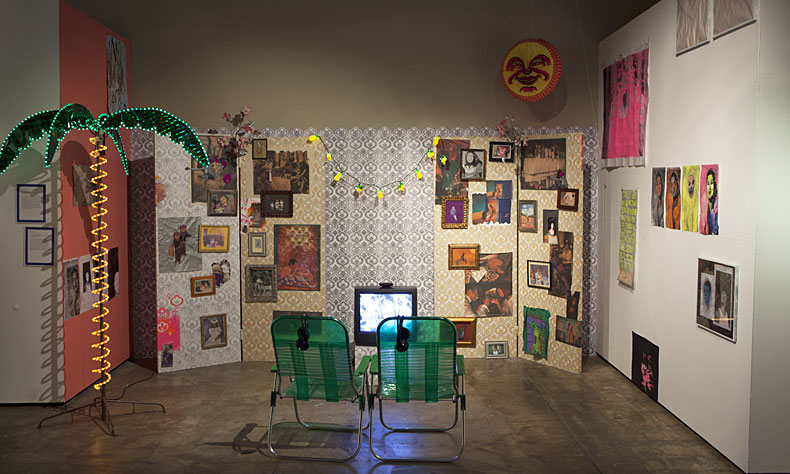 Installation mit zwei Campingstühlen vor Wänden mit gemusterter Tapete und vielen Bildern und einer Palme aus Lichtschläuchen