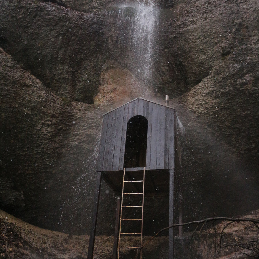 eine Holzhütte auf Stelzen steht an einem Felsen unter einem Wasserfall